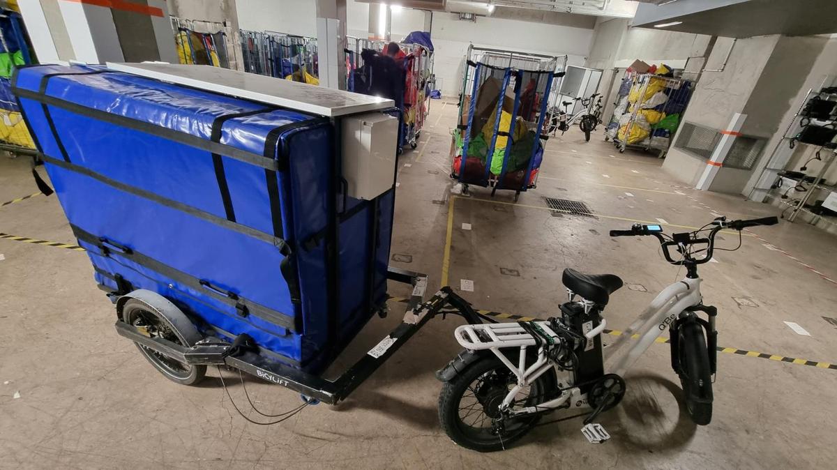 Conteneur isotherme bleu, équipé d'un panneau solaire et d'un boîtier blanc, monté sur une remorque BicyLift attelée à un vélo électrique.